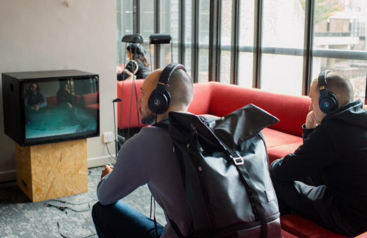 Two audience members sit on a sofa in the Barbican Centre foyer. They are wearing headphones and watching a film of Bluey in a bed on a thick old school box telly.